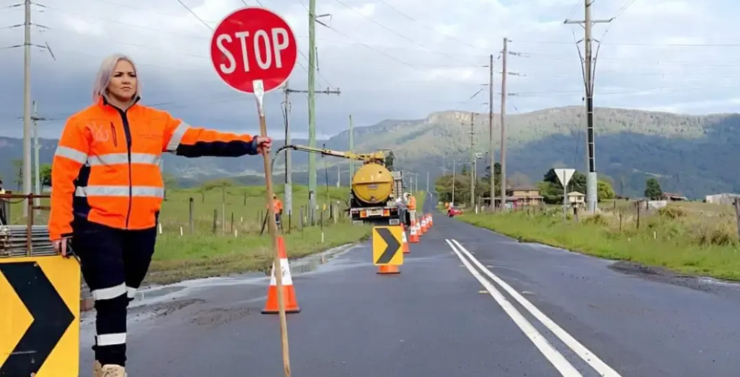 A Crown traffic controller holding a stop/slow bat beside a road